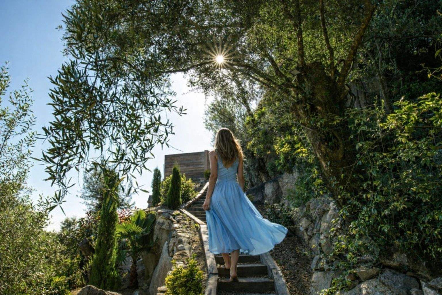 Femme en robe bleue montant les escaliers vers une villa de luxe méditerranéenne entourée de végétation verdoyante